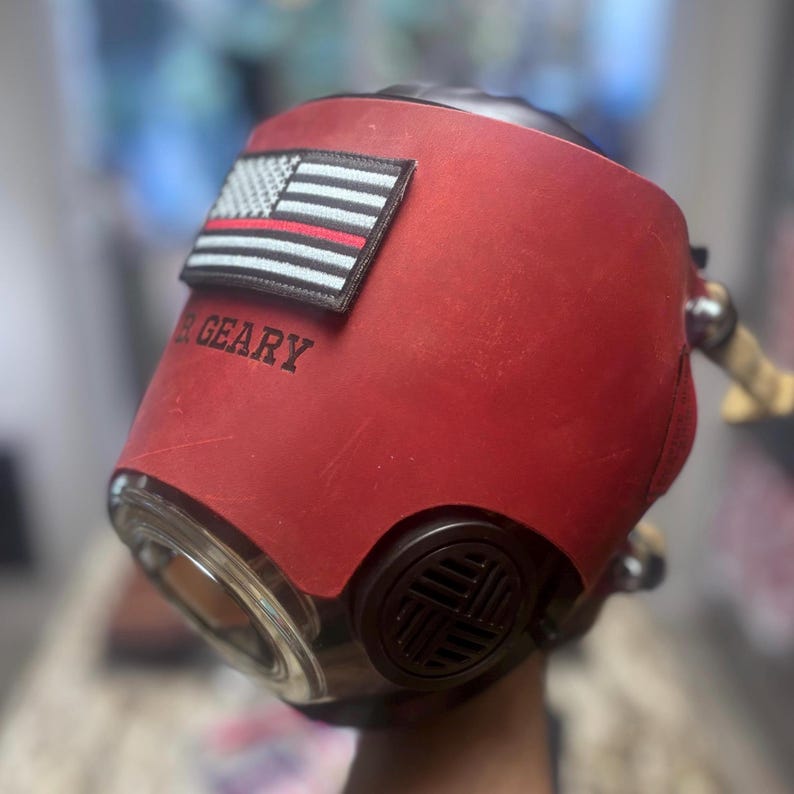 May include: A red leather protective mask featuring a black and white American flag patch with a thin red line. The name R. GEARY is embossed on the side. The mask includes a circular clear lens and a dark brown circular ventilation filter component.