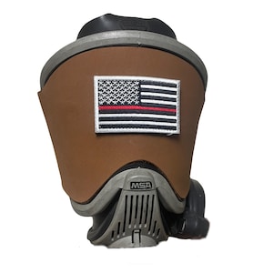 May include: A brown leather cover for an MSA respirator mask featuring a rectangular embroidered patch of the American flag with a thin red horizontal line across the center. The cover is shaped to fit the front of the gray respirator unit.