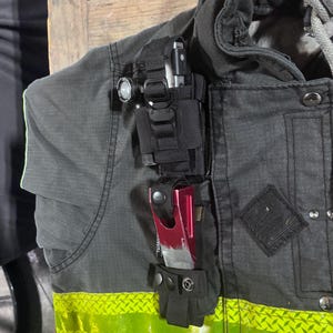 May include: A black tactical gear mount attached to a dark gray jacket with a high-visibility yellow reflective strip. The mount holds a small black flashlight at the top and a metallic red tool with a rectangular cutout secured by a snap button.