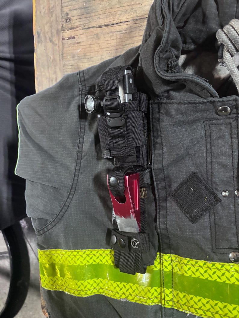 May include: A black tactical gear mount attached to a dark gray jacket with a high-visibility yellow reflective strip. The mount holds a small black flashlight at the top and a metallic red tool with a rectangular cutout secured by a snap button.