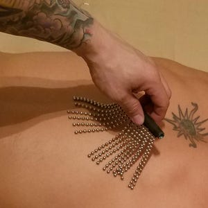 Könnte beinhalten: Ein Massage-Werkzeug mit einem schwarzen Griff und mehreren Strängen kleiner, silberfarbener Perlen. Das Werkzeug wird auf dem Rücken einer Person in der Nähe eines Sonnentattoos verwendet.