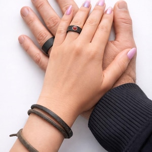 May include: Two black rings, one with a red stone, are displayed on hands. A pair of dark brown leather bracelets are also visible. The image is set against a white background, with a black long-sleeve shirt in the corner.