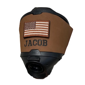 Puede incluir: Un respirador negro y marrón con el nombre "JACOB" y un parche de la bandera estadounidense. El respirador tiene una abertura circular y la marca "Dräger" es visible.