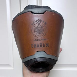 May include: A brown leather fire helmet band featuring a black embossed logo of the International Association of Fire Fighters, the text LOCAL 1595, and the name GRAHAM printed in black. The band is attached to a dark gray fire helmet base.