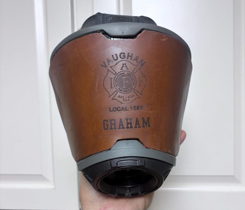 May include: A brown leather fire helmet band featuring a black embossed logo of the International Association of Fire Fighters, the text LOCAL 1595, and the name GRAHAM printed in black. The band is attached to a dark gray fire helmet base.