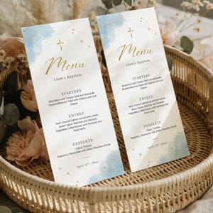 May include: Two baptism menus with a blue watercolour design and gold text, featuring a cross and the name "Liam's Baptism." The menus list starters, entree, and dessert options. They are displayed in a woven basket with floral accents.