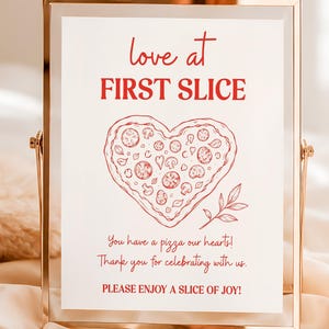Könnte beinhalten: Ein vertikales rechteckiges Schild in einem goldfarbenen Metallrahmen mit rotem Text, der love at FIRST SLICE lautet, einer Strichzeichnung einer herzförmigen Pizza und dem Text You have a pizza our hearts! Thank you for celebrating with us. PLEASE ENJOY A SLICE OF JOY!