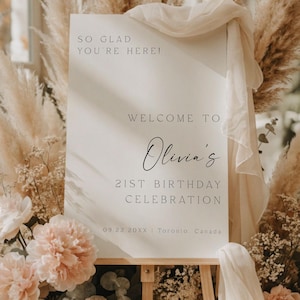 May include: A white welcome sign on a wooden easel, surrounded by pampas grass and peach-colored flowers. The sign reads "SO GLAD YOU'RE HERE!" and "WELCOME TO Olivia's 21ST BIRTHDAY CELEBRATION".