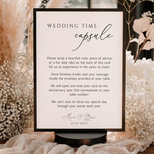 May include: A rectangular sign in a black frame with the text Wedding Time Capsule. The sign includes instructions for guests to write notes for an anniversary, featuring the names Maria & Bren and the date 07.07.2029 in black lettering.