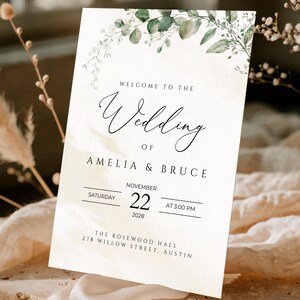 Może przedstawiać: Prostokątna tablica powitalna na ślub z zielonym akwarelowym łukiem z eukaliptusa u góry. Tekst brzmi: Welcome to the Wedding of Amelia & Bruce, Saturday November 22, 2028 at 3:00 PM, The Rosewood Hall, 278 Willow Street, Austin.