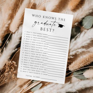 以下が含まれることがあります： 「WHO KNOWS THE graduate BEST?」と書かれた黒い文字の白いカード。卒業帽子のグラフィックが付いています。カードには空欄の質問があります。カードは乾燥した植物に囲まれています。