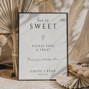 Peut inclure: Un panneau encadré avec le texte "love is SWEET" et "PLEASE TAKE A TREAT". Le panneau comprend également les noms "CHLOE + RYAN" et la date "09.06.2028". Le panneau est entouré de plantes séchées et de tissu.