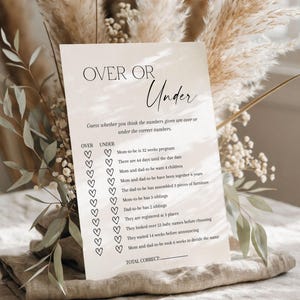 May include: A beige card with the text "OVER OR Under" and a list of questions for a baby shower game. The card is decorated with heart-shaped check boxes and is surrounded by dried flowers and greenery.