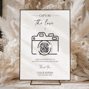 May include: A rectangular white sign with black text and a camera icon featuring a QR code. The text reads: CAPTURE the love, SCAN TO UPLOAD, SHARE YOUR PHOTO WITH US, SCAN WITH YOUR PHONE AND UPLOAD, Thank You, CHLOE & RYAN, 14TH NOVEMBER 20XX.