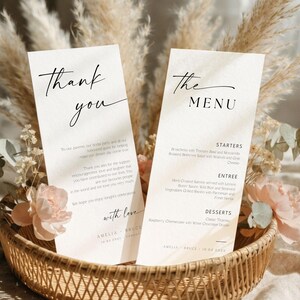 May include: Two elegant wedding menu cards and a thank you card in a woven basket, adorned with dried flowers and pink blossoms. The cards feature handwritten-style text, including "Thank You" and "The Menu."
