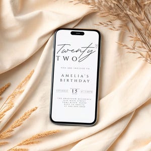 Könnte beinhalten: Ein Smartphone zeigt eine digitale Einladung zum Geburtstag von Amelia. Die Einladung enthält die Worte "Twenty Two" in eleganter Schrift sowie Details zur Veranstaltung. Das Telefon liegt auf einem beigen Stoff mit getrockneten Weizenähren.