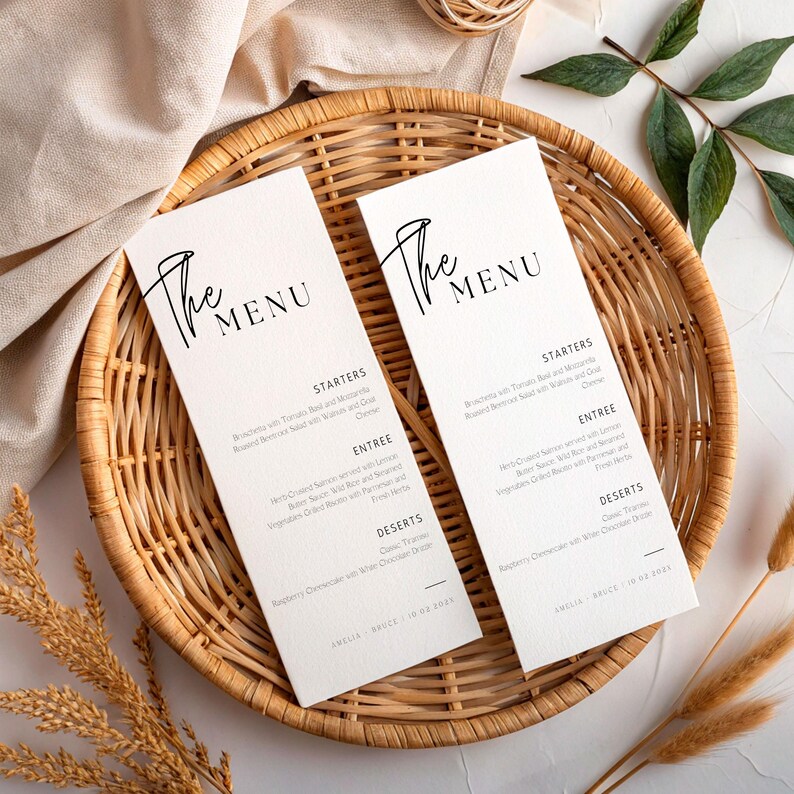 May include: Two rectangular white menu cards with the text "The Menu" in elegant black script, displayed in a woven wicker basket. The menu cards list starters, entree, and dessert options. The cards are surrounded by decorative wheat and greenery.