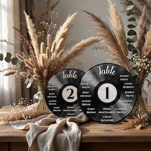 May include: Two black vinyl record-shaped table signs with white text and numbers. The signs are labeled "Table 1" and "Table 2" with guest names around the edges. The background includes dried flowers and a wooden table.