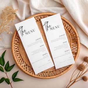 May include: Two white menu cards with black text, displayed on a round woven tray. The cards read "The Menu" in elegant script. Menu items are listed below, including starters, entree, and desserts. The tray is set on a neutral-toned surface.