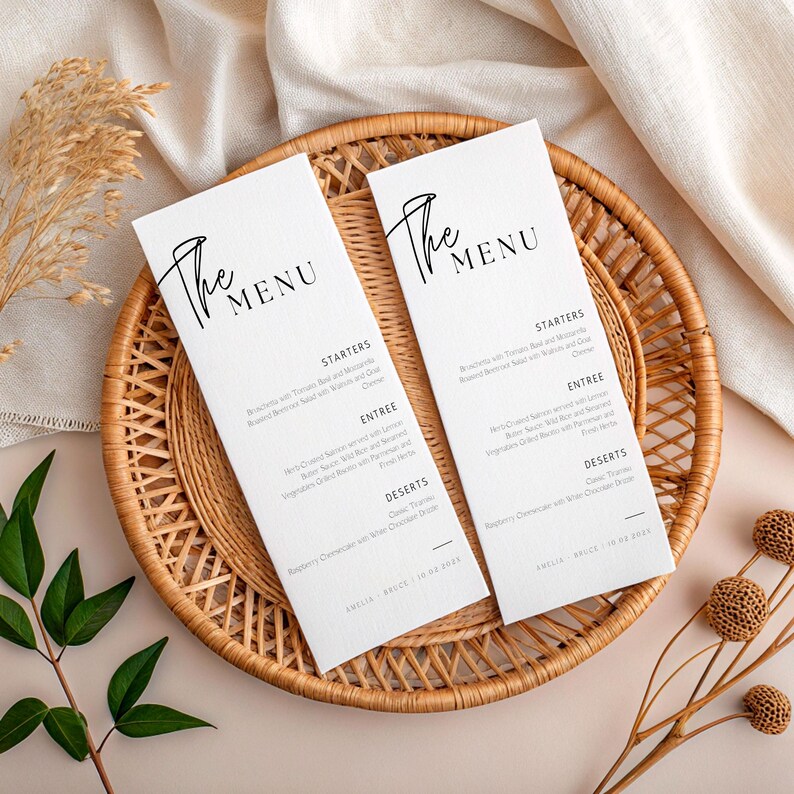 May include: Two white menu cards with black text, displayed on a round woven tray. The cards read "The Menu" in elegant script. Menu items are listed below, including starters, entree, and desserts. The tray is set on a neutral-toned surface.