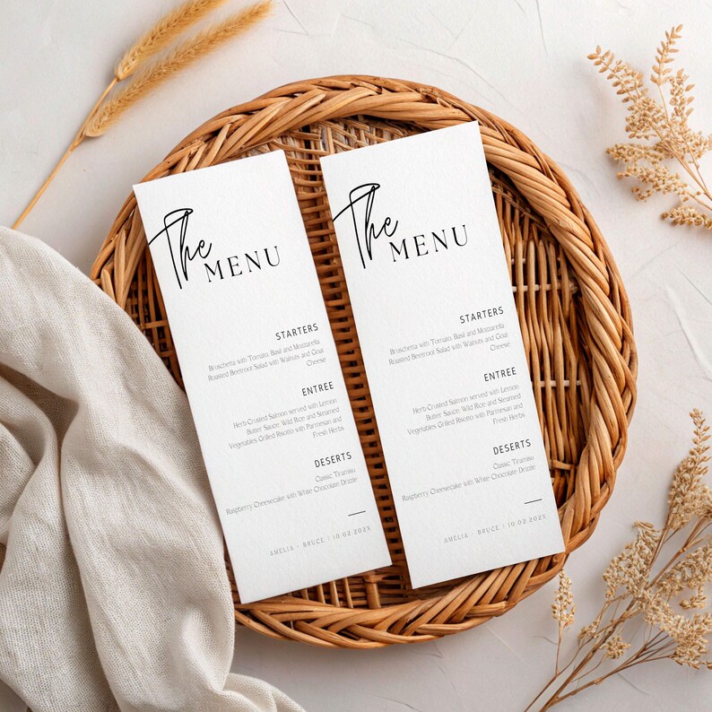 May include: Two white menu cards with black text, featuring the words "The Menu" in elegant script. The cards are placed on a woven wicker tray, with a linen cloth and dried wheat stalks in the background. The menu lists starters, entree, and desserts.
