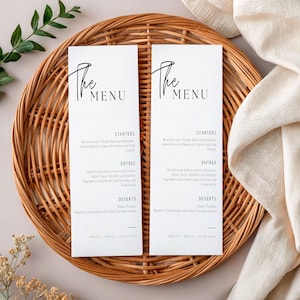 May include: Two white menu cards with black script, reading "The Menu." The cards list starters, entree, and dessert options. The cards are on a woven wicker tray with a neutral background. The menu is for Amelia and Bruce's wedding.