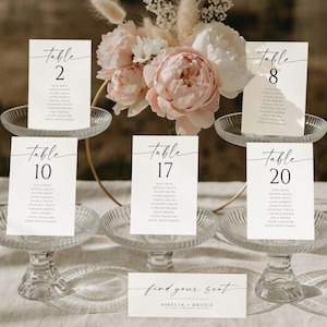 Puede incluir: Elegantes tarjetas con números de mesa para bodas con un diseño minimalista. Cada tarjeta presenta un número de mesa en una fuente elegante, acompañado de los nombres de los invitados. Las tarjetas se exhiben sobre soportes de vidrio, complementadas con arreglos florales.
