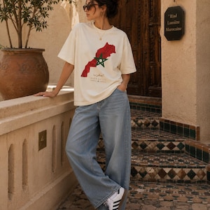 May include: An off-white short-sleeved t-shirt featuring a red map of Morocco with a green star in the center, accompanied by the text "المغرب" and "MOROCCO" in black, paired with light blue wide-leg denim jeans and white sneakers with black stripes.