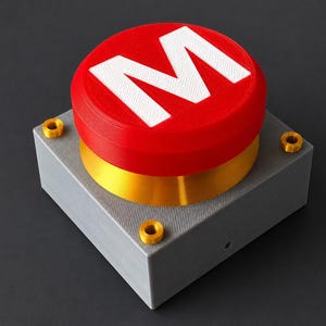 Può includere: Un grande pulsante rosso con una "M" bianca in cima, posto su una base quadrata grigia con accenti dorati. Il pulsante è di un rosso vibrante e la base ha una superficie strutturata. Quattro viti color oro fissano il pulsante alla base.