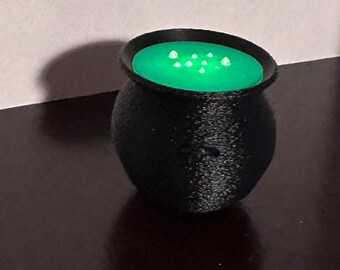 May include: A small, black cauldron with a textured surface. The cauldron has a green interior with several white, glowing dots, suggesting a bubbling potion. The cauldron is likely a decorative item or a prop.