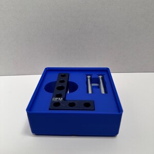 May include: A blue box contains black metal brackets and silver screws. The brackets are L-shaped with multiple holes. The text "DFM" is visible on one of the brackets. The box is on a white surface.