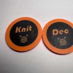 May include: Two orange and black circular craft tokens. One token reads "Knit" with a yarn ball graphic. The other reads "Dec" with a yarn ball graphic. These tokens are likely used for knitting or crafting projects.