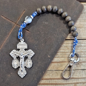 May include: A religious keyring featuring a silver-toned crucifix with the inscription JESUS NAZARENUS REX JUDAEORUM, flanked by two small oval medallions. The crucifix is attached to a blue cord with a white bead and a series of dark wooden beads.