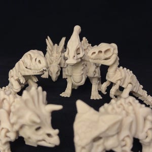 Puede incluir: Una colección de modelos de esqueletos de dinosaurios de color blanquecino, incluyendo un Tyrannosaurus Rex, Triceratops, y otros. Los modelos están dispuestos en círculo, mostrando sus estructuras esqueléticas y detalles. El fondo es negro liso.