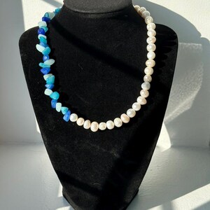 May include: A necklace displayed on a black mannequin. The necklace features a combination of white pearls and blue-toned stones. The stones transition from dark blue to light blue. The pearls are white and round.