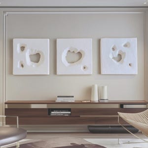 May include: Three white square wall art pieces with irregular, organic-shaped cutouts. The art is mounted above a dark wood cabinet with books and decorative vases. The room has a modern, minimalist aesthetic.