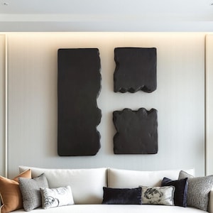 Pode incluir: Três peças de arte de parede abstratas pretas com bordas texturizadas são montadas em uma parede cinza claro acima de um sofá branco com almofadas decorativas. As peças de arte têm formas retangulares e quadradas.