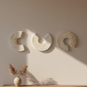 Op de afbeelding: Drie abstracte wandkunstwerken in crème- en beigetinten. Elk heeft een unieke, onvolledige cirkelvorm. Daaronder staat een kleine beige vaas met gedroogd siergras.