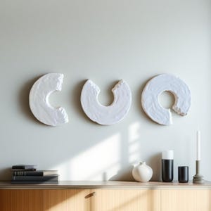 Peut inclure: Trois sculptures murales en céramique blanche, chacune en forme de cercle partiel avec un bord texturé. Les sculptures sont montées sur un mur gris clair au-dessus d'un meuble en bois. Des objets décoratifs, dont une bougie et des vases, sont sur le meuble.