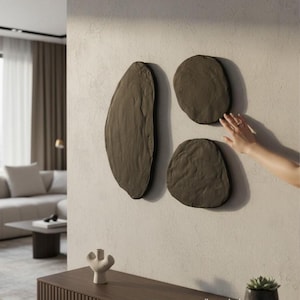 Op de afbeelding: Drie donkergrijze, getextureerde wandkunstwerken in organische vormen zijn bevestigd aan een lichtgrijze muur. De vormen omvatten een langwerpig ovaal en twee afgeronde vormen. Een hand reikt naar de kunst.