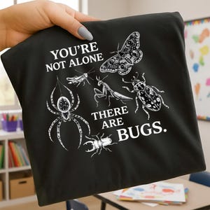May include: Black t-shirt featuring a variety of bug illustrations in white, including a spider, moth, and beetle. The text "YOU'RE NOT ALONE THERE ARE BUGS" is printed above and below the bug graphics.