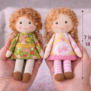 Pode incluir: Duas bonecas de tecido feitas à mão, cada uma com 18 cm de altura, com cabelo ruivo encaracolado e bochechas rosadas. Uma boneca usa um vestido floral verde, a outra um vestido floral amarelo. Ambas têm detalhes brancos e castanhos.