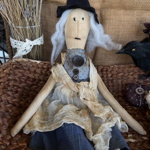 May include: A handcrafted fabric witch doll wearing a black pointed hat, white hair, and a blue and cream-coloured dress. The doll has button details and is seated on a woven basket. A decorative crow and lavender bouquet are in the background.