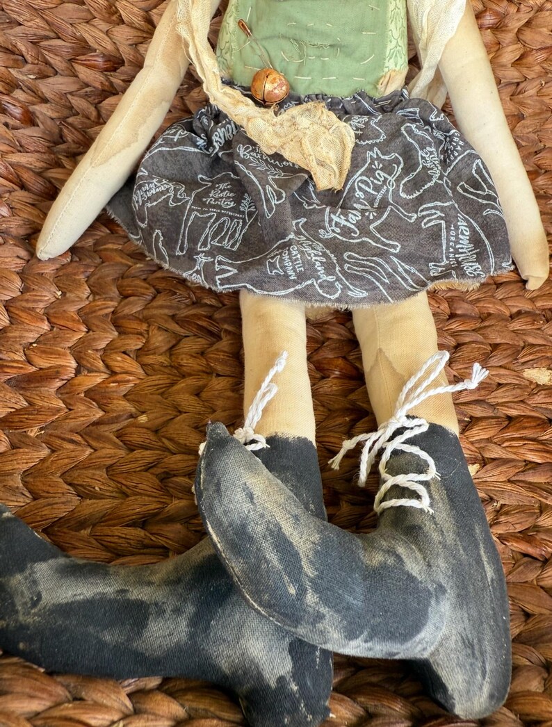 Puede incluir: Mu&ntilde;eca de tela hecha a mano con un delantal verde, una falda con estampado de animales de granja y botas negras con cordones blancos. La mu&ntilde;eca tiene brazos y piernas de color beige y cabeza y torso beige.