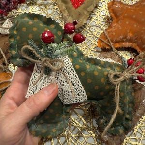May include: A handmade Christmas ornament featuring a green fabric angel with gold polka dots, adorned with red berries and a lace collar. Other ornaments include a heart and star shape, all with rustic, homespun details.