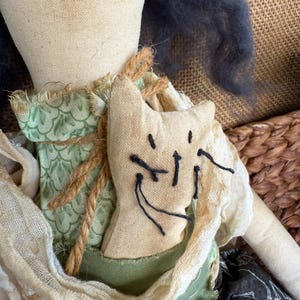Puede incluir: Una mu&ntilde;eca hecha a mano con una aplicaci&oacute;n en forma de gato. La mu&ntilde;eca tiene un cuerpo de tela beige, ropa verde y beige y cabello azul oscuro. La aplicaci&oacute;n de gato est&aacute; cosida con hilo negro y la mu&ntilde;eca est&aacute; adornada con cuerda.