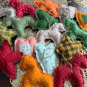 Op de afbeelding: Een verzameling handgemaakte stoffen ornamenten in verschillende vormen en patronen. De ornamenten omvatten paarden, sterren en harten in rode, oranje, groene en bloemenprints. Elk ornament heeft een touwtje om op te hangen.