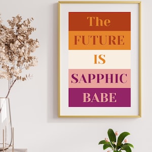 May include: Framed art print with the text "The FUTURE IS SAPPHIC BABE" in a bold, sans-serif font. The text is layered over horizontal bands of orange, cream, pink, and purple. The frame is gold.