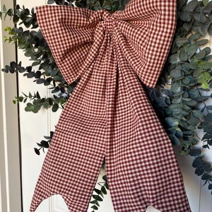 May include: A large, decorative bow made of red and white checkered fabric. The bow is attached to a green eucalyptus wreath, creating a rustic and charming aesthetic. The bow's tails have pointed ends.