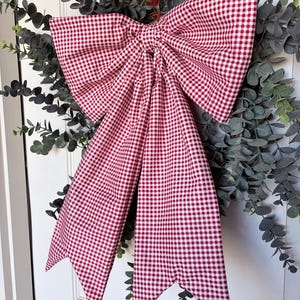 May include: A large, decorative bow made of red and white gingham fabric. The bow is attached to a wreath of green eucalyptus leaves. The bow's tails have pointed ends, and the bow is centered on a white door.